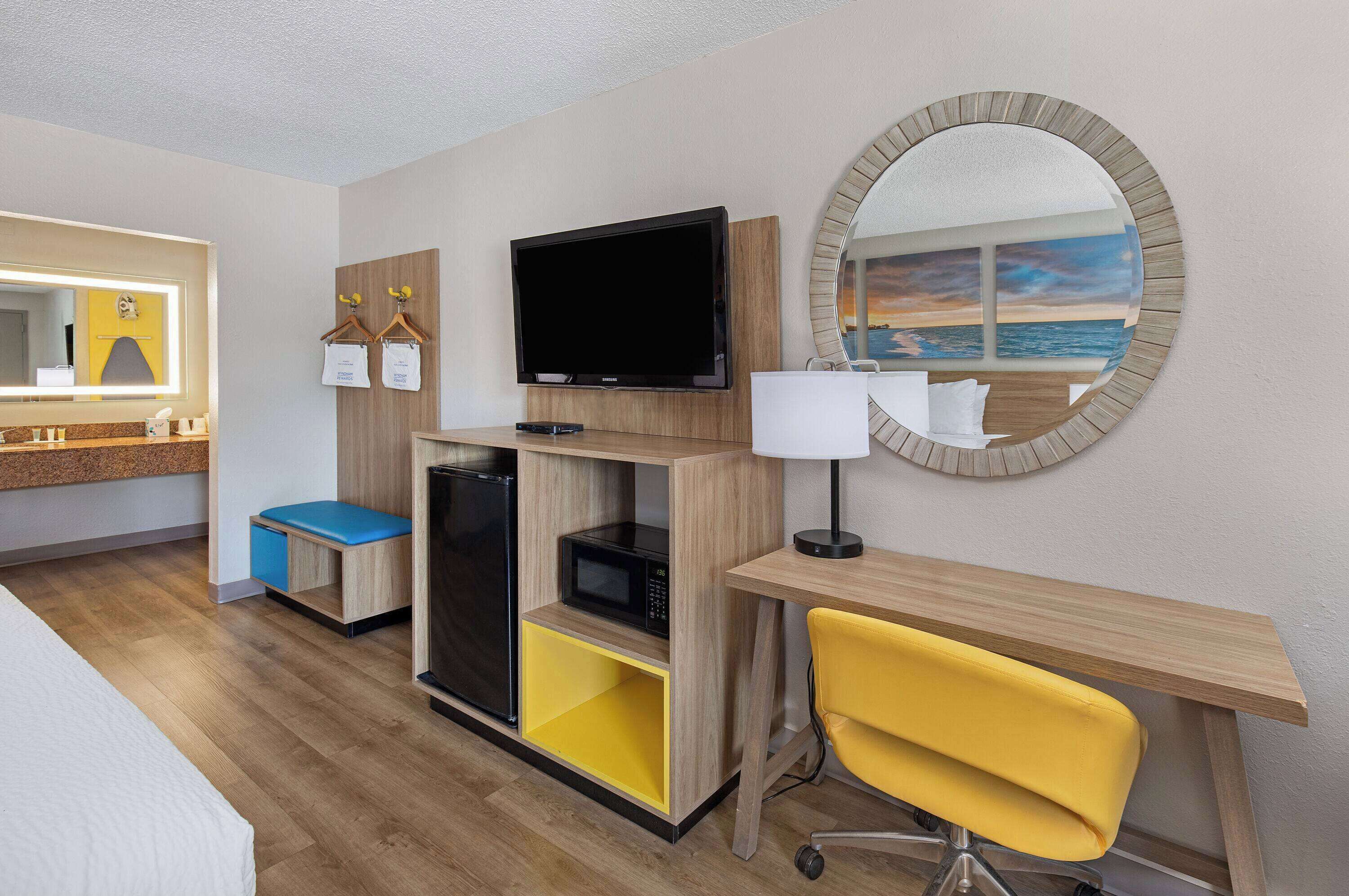 Habitación de hotel con cama, escritorio con silla, televisor, microondas, refrigerador pequeño y espejo con vista a la playa.