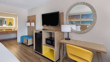 A hotel room with a bed, a desk with a chair, a television, a microwave, a small refrigerator, and a mirror with a beach view.