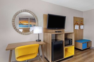 A compact room with a wooden desk, a yellow chair, a flat-screen TV, a refrigerator, a microwave, and a small seating area with a blue cushion.