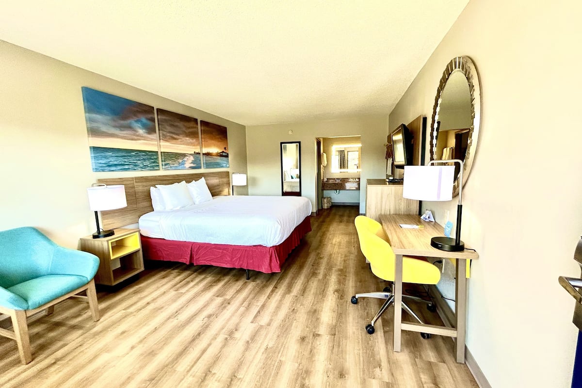 A hotel room with a bed, a desk, a chair, a mirror, and a painting of a beach scene.