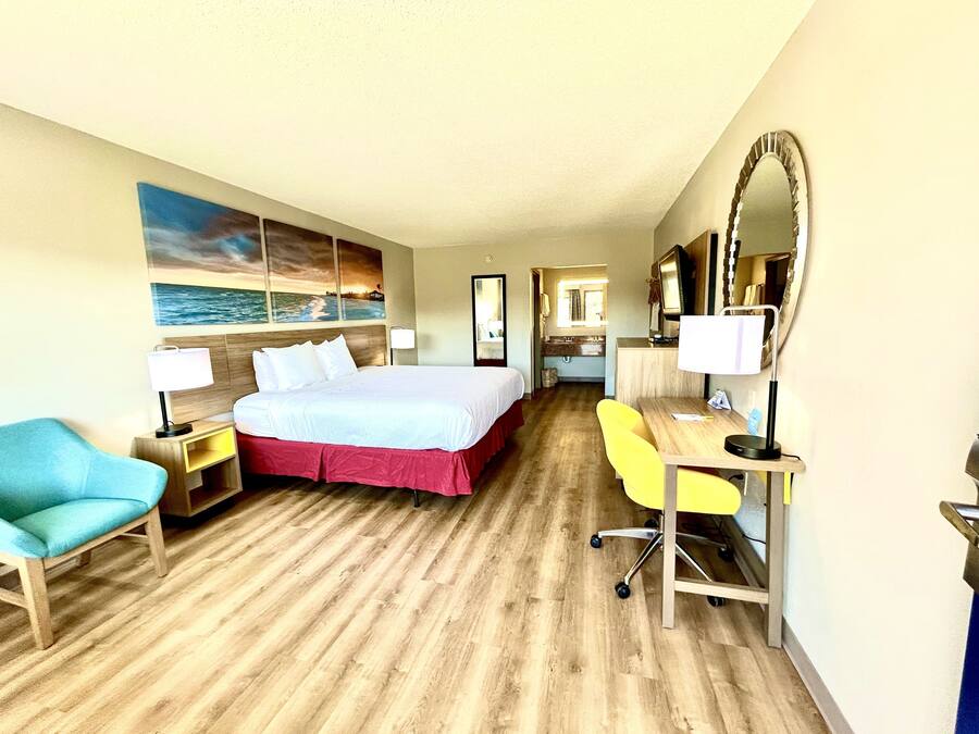 A hotel room with a bed, a desk, a chair, a mirror, and a painting of a beach scene.