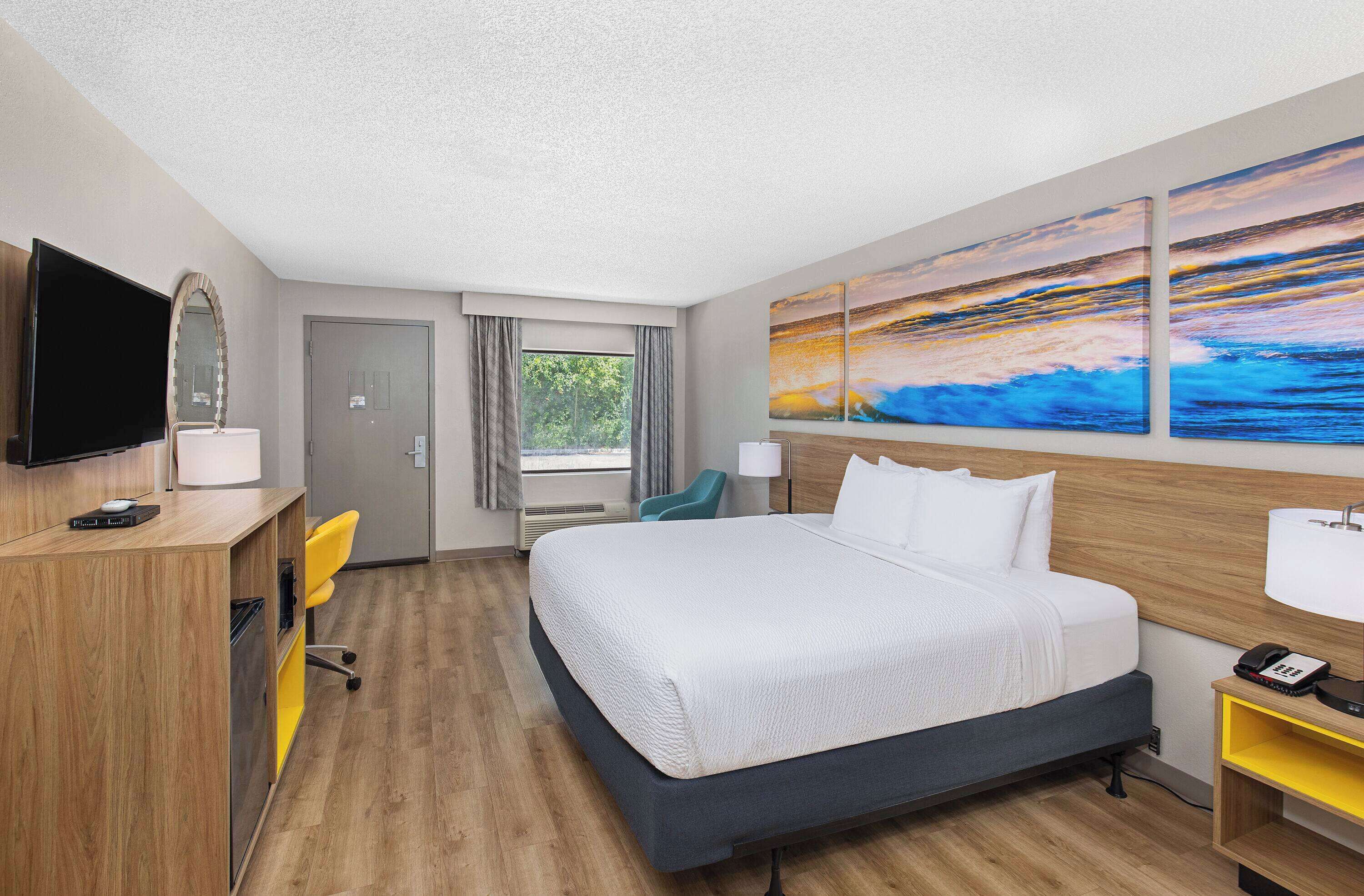 A hotel room with a bed, a desk with a TV, a chair, a desk lamp, a bedside table with a lamp, and artwork on the wall.