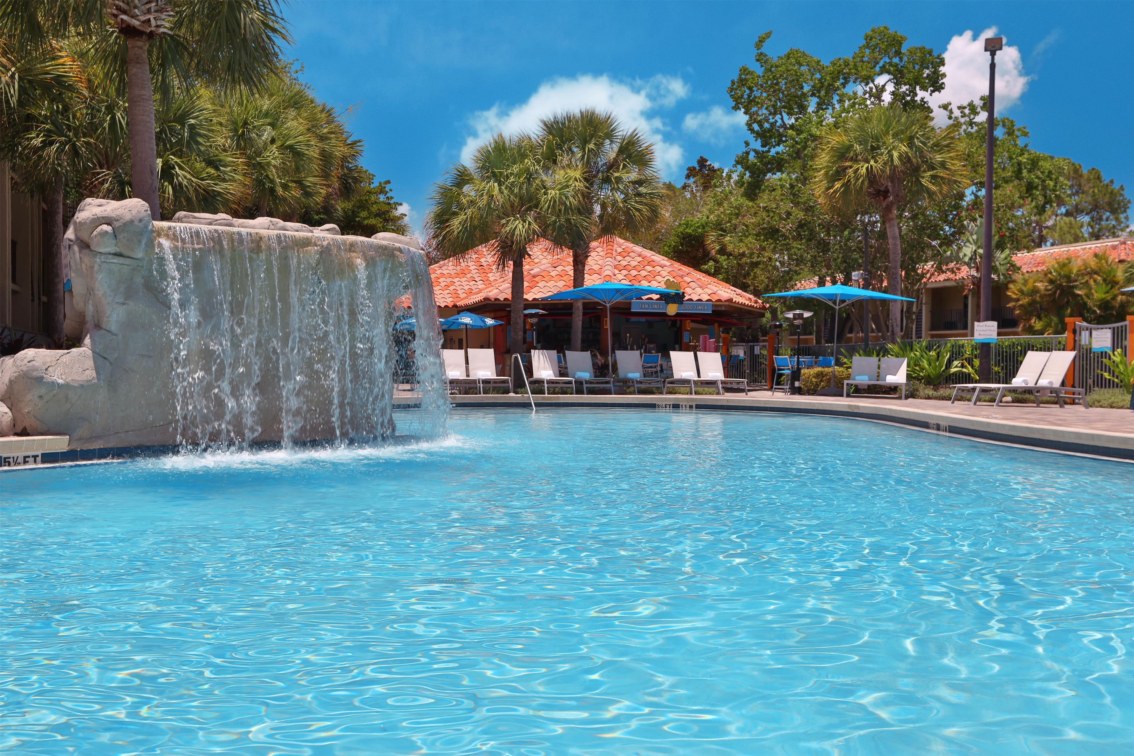 A swimming pool with a waterfall feature, lounge chairs, and a building with a red-tiled roof in the background.