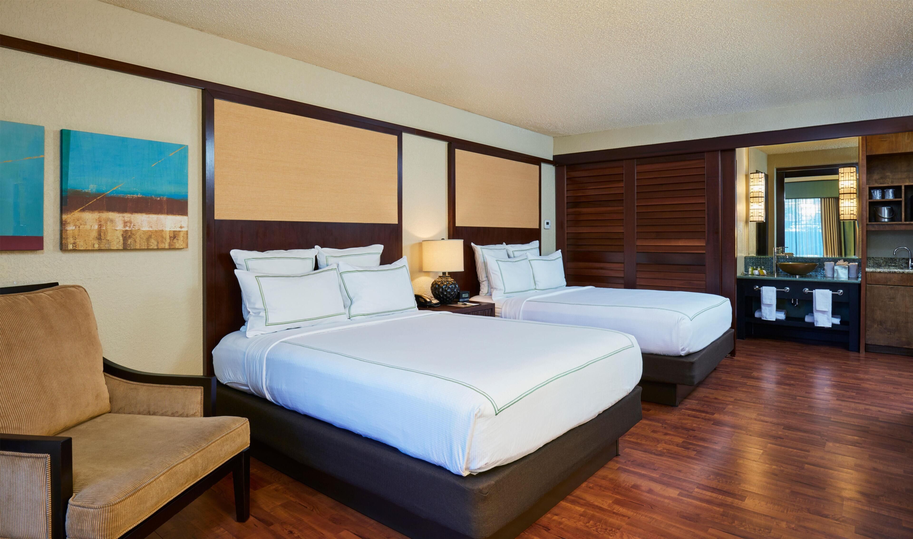A hotel room with two beds, a chair, a desk, a mirror, and a bathroom with a sink and vanity.