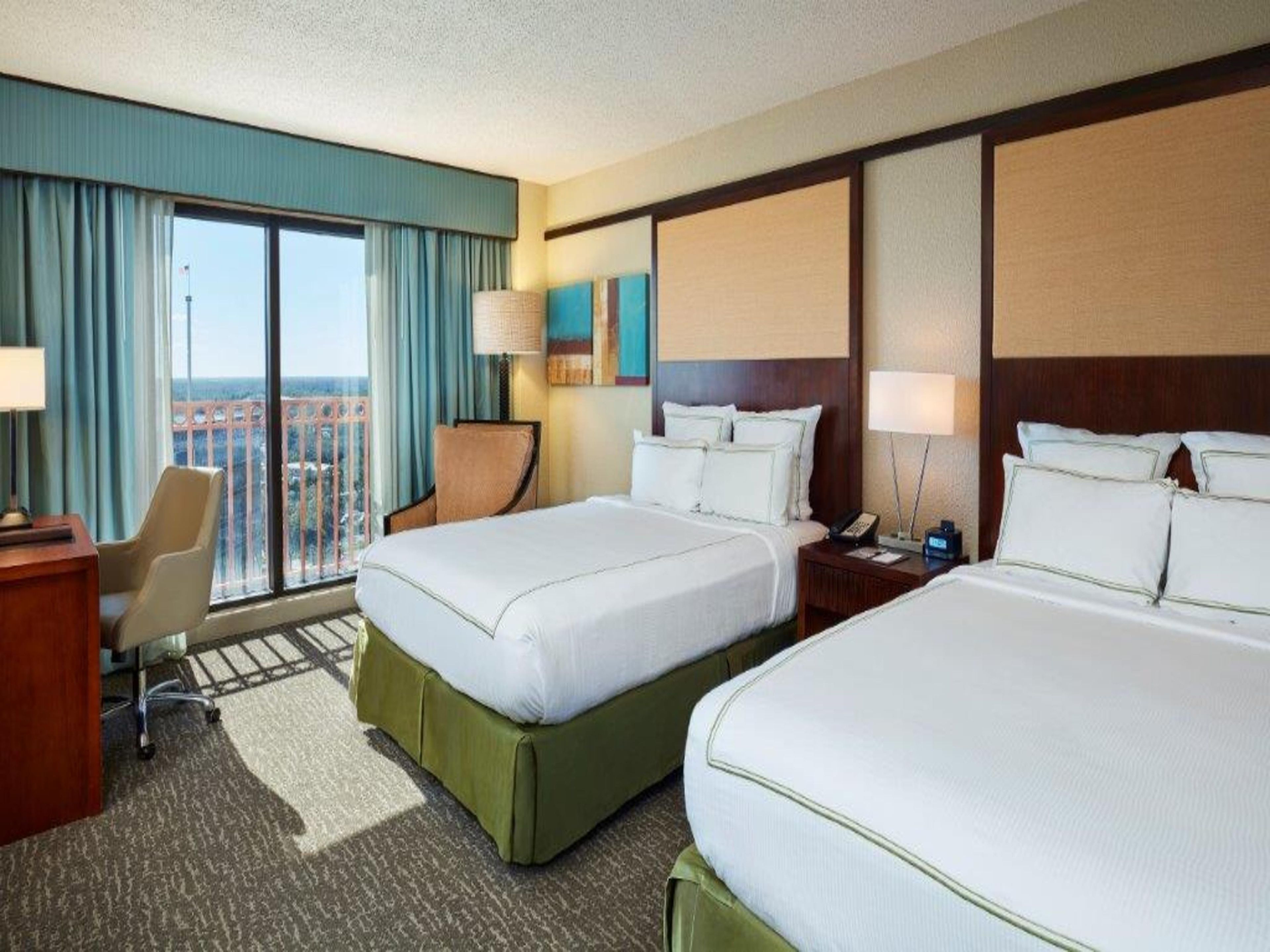 A hotel room with two beds, a desk, a chair, and a view of the city.