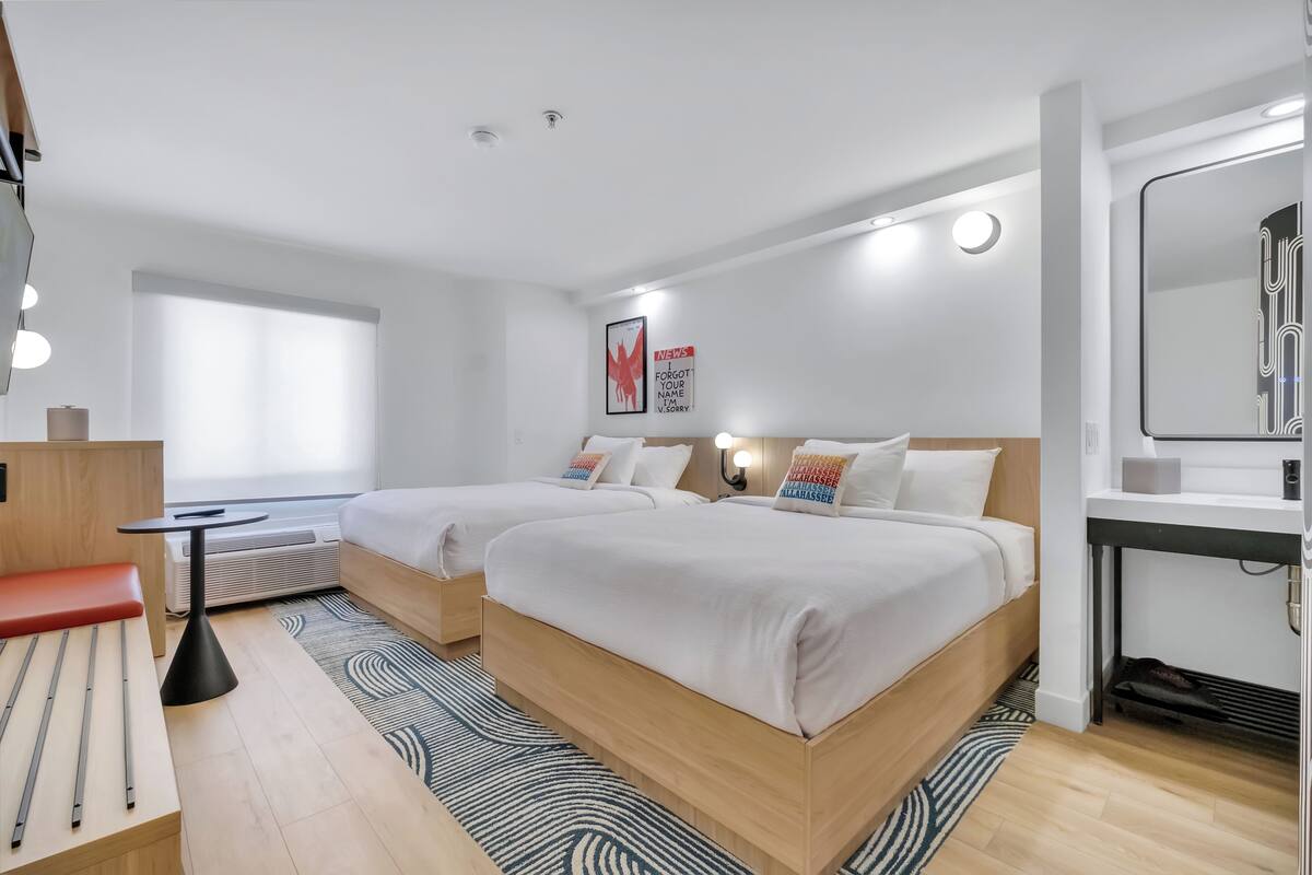 A modern hotel room with two beds, a small desk, a chair, a mirror, and a window with blinds.