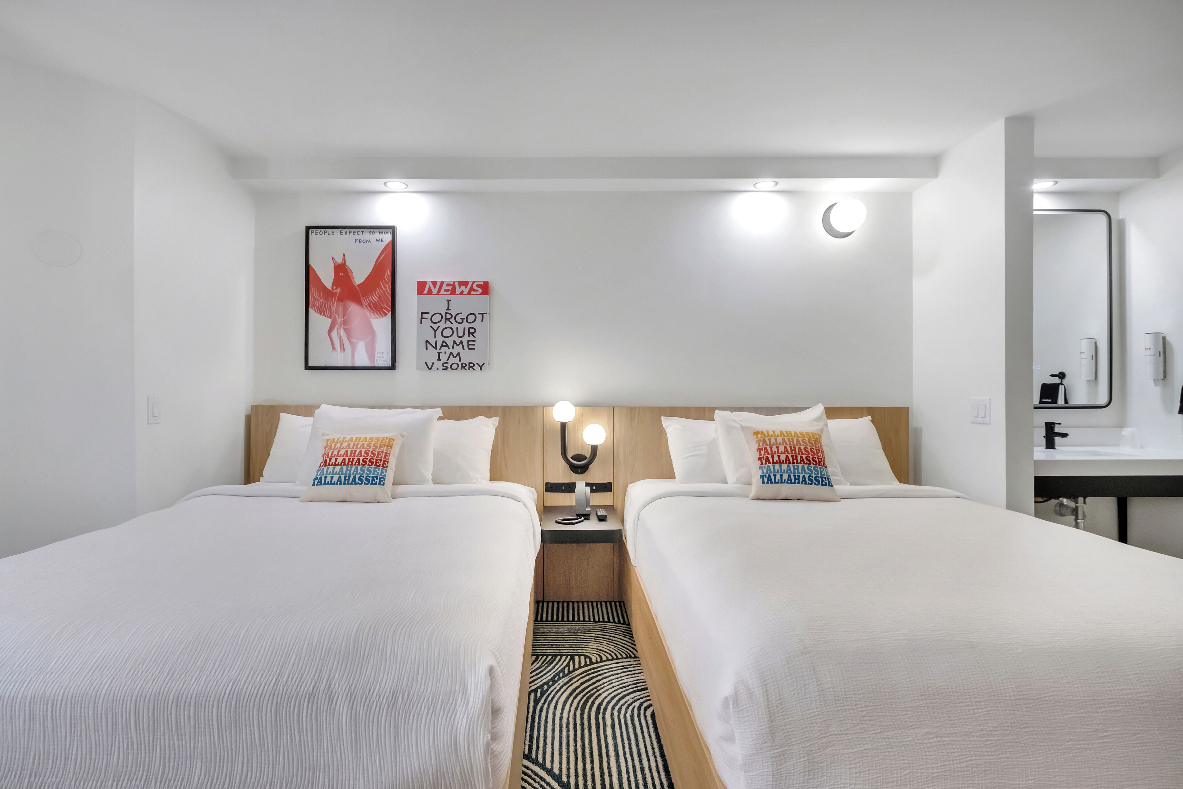 Une chambre d’hôtel avec deux lits, une table de chevet avec une lampe, un téléviseur fixé au mur et une salle de bain visible à travers une porte ouverte.
