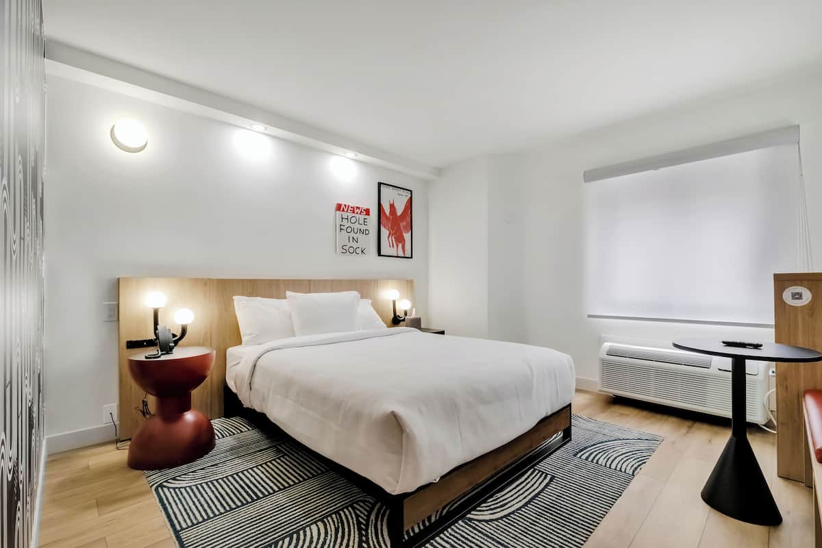 A modern hotel room with a large bed, bedside lamps, a small table, and a window with blinds.