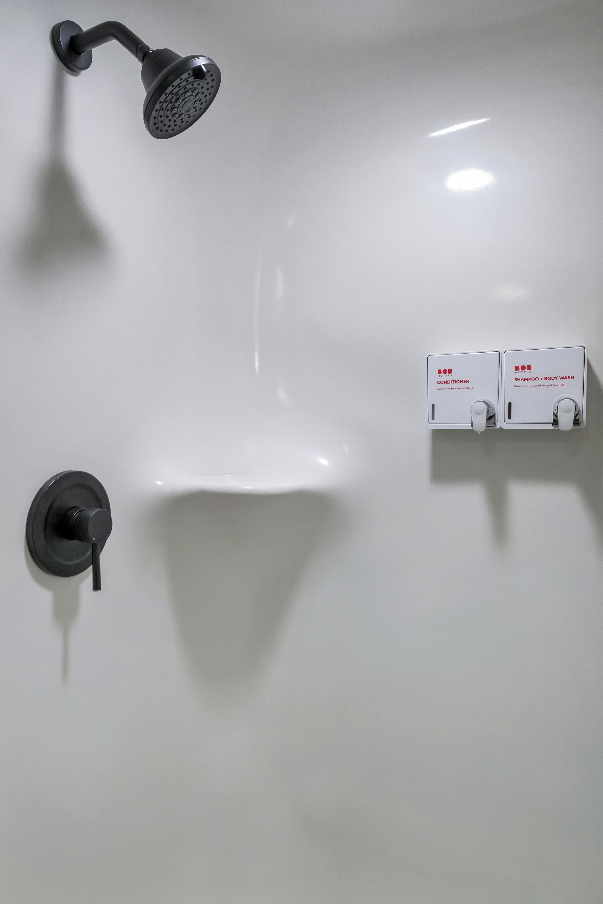 Un baño con un lavamanos blanco, un cabezal de ducha manual negro y dos dispensadores de jabón blancos etiquetados como 'Acondicionador' y 'Champú + Gel de Baño'.