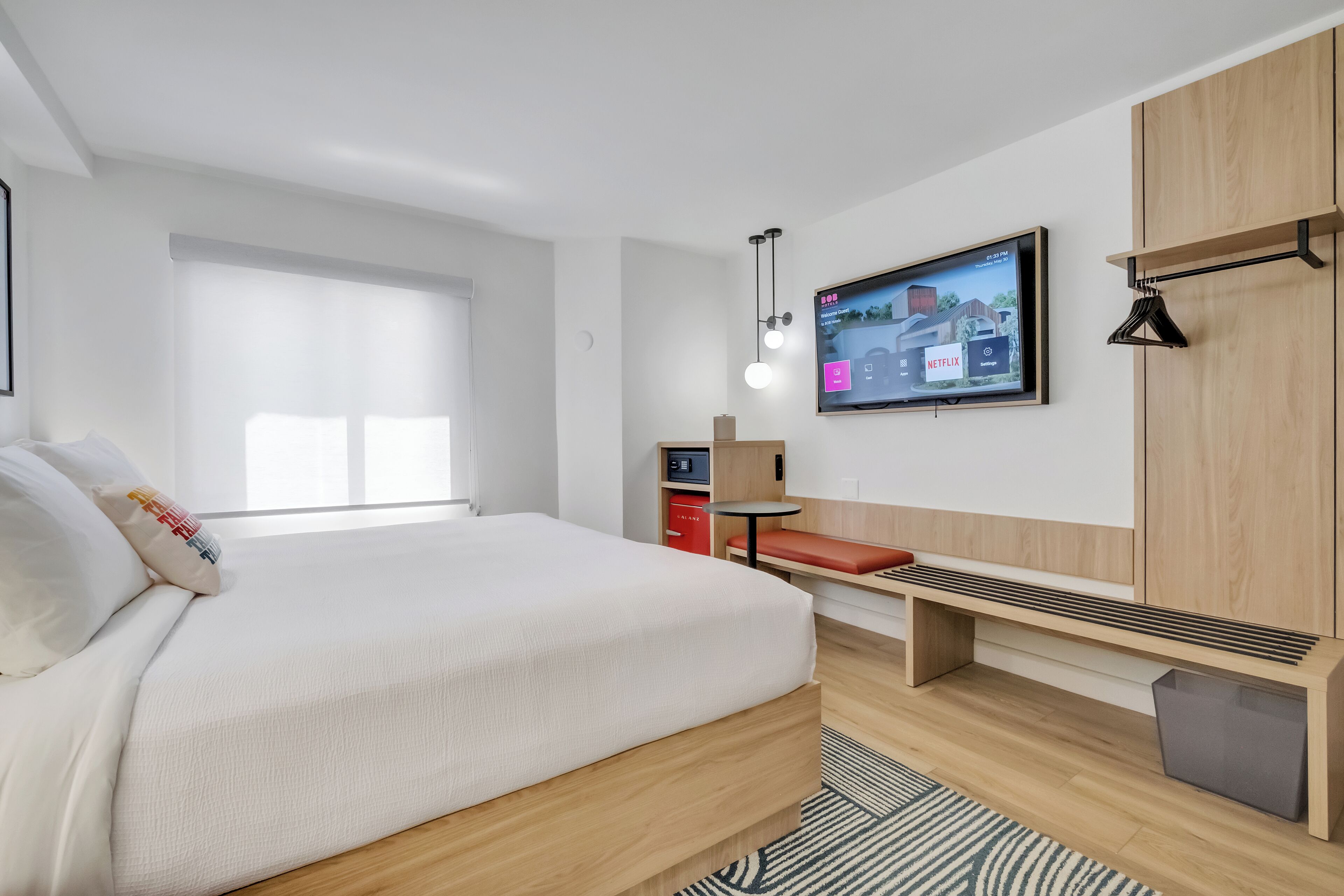 A modern hotel room with a large bed, a TV mounted on the wall, a small desk, and a bench.
