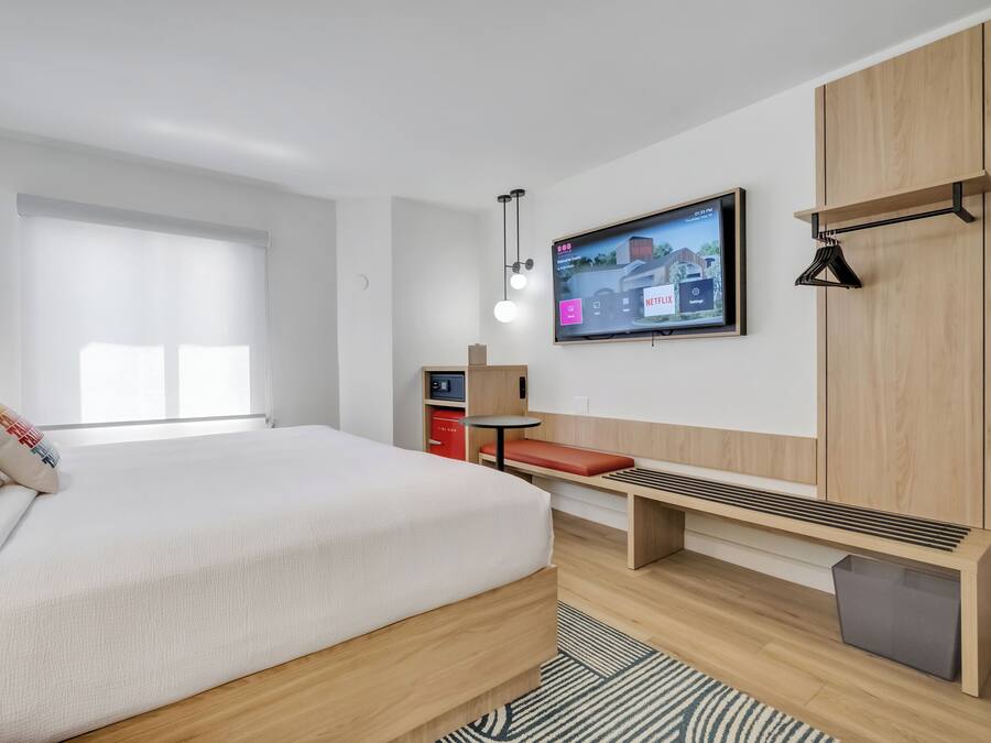 A modern hotel room with a large bed, a TV mounted on the wall, a small desk, and a bench.