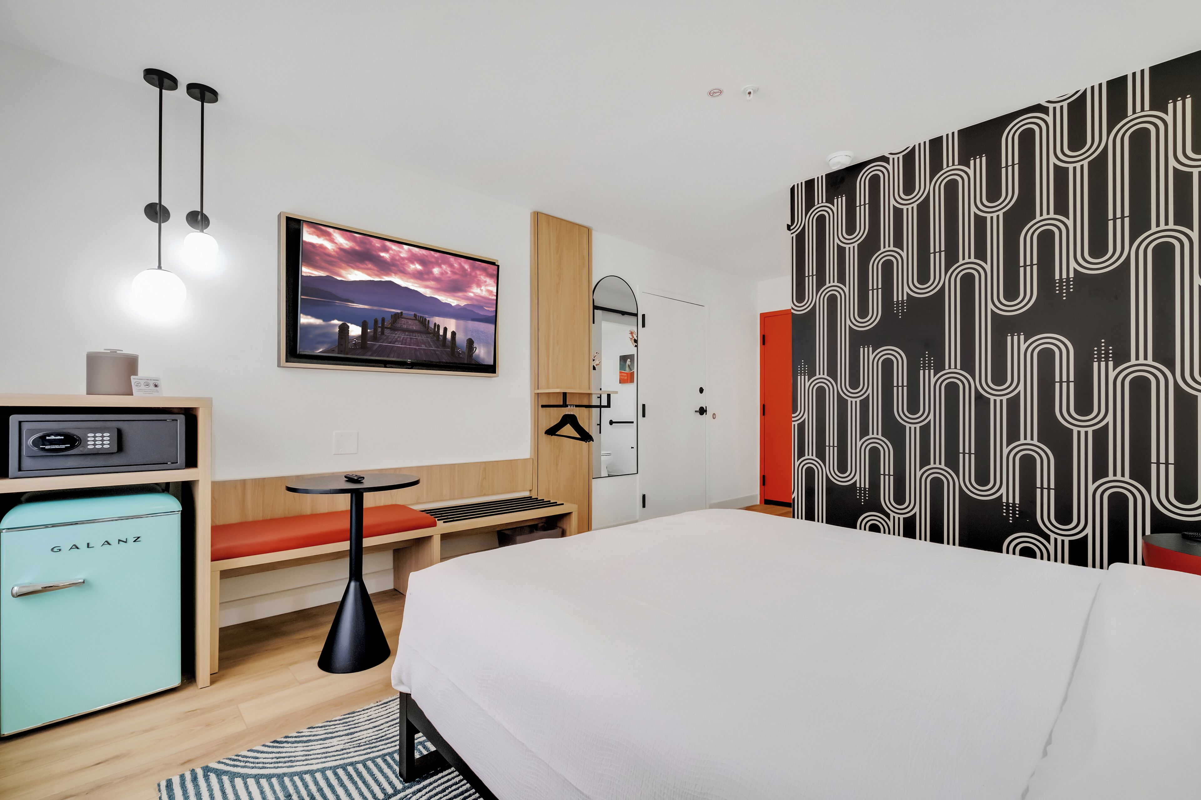 Een moderne hotelkamer met een bed, een bureau, een kleine koelkast, een hanglamp en een spiegel aan de muur.