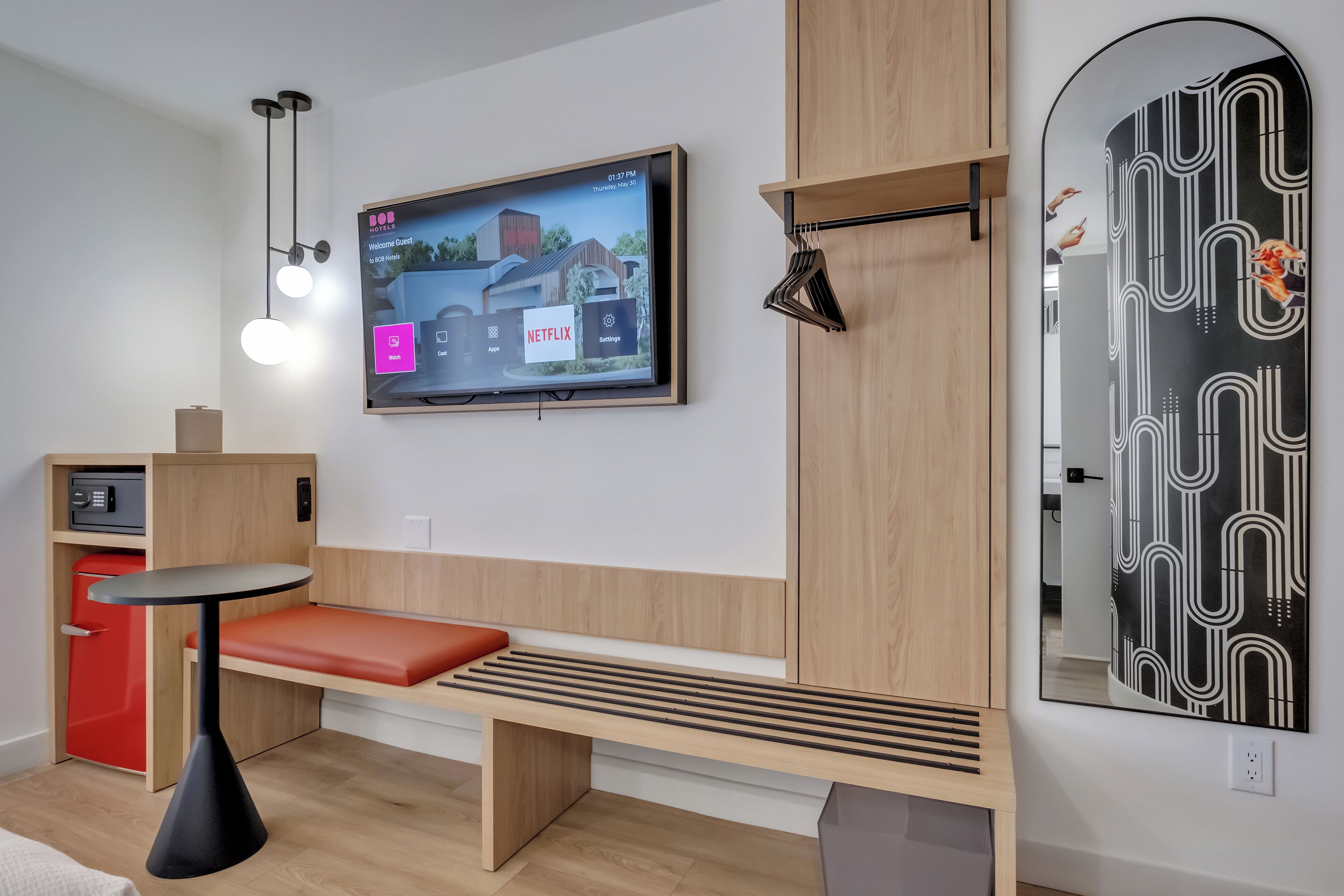 Een moderne hotelkamer met een houten bankje, een klein tafeltje, een aan de muur bevestigde televisie en een rode minibars.
