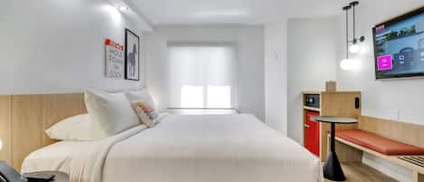 A modern hotel room with a large bed, a bedside table with a phone, a small desk with a chair, a TV mounted on the wall, and a window with blinds.