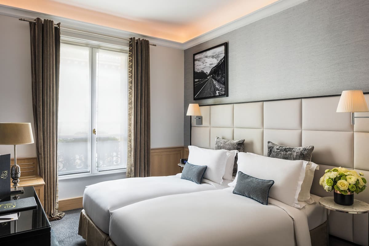 Ett hotellrum med två sängar, ett stort fönster med gardiner, en sänggavel med en tavla och ett litet bord med en lampa och blommor.