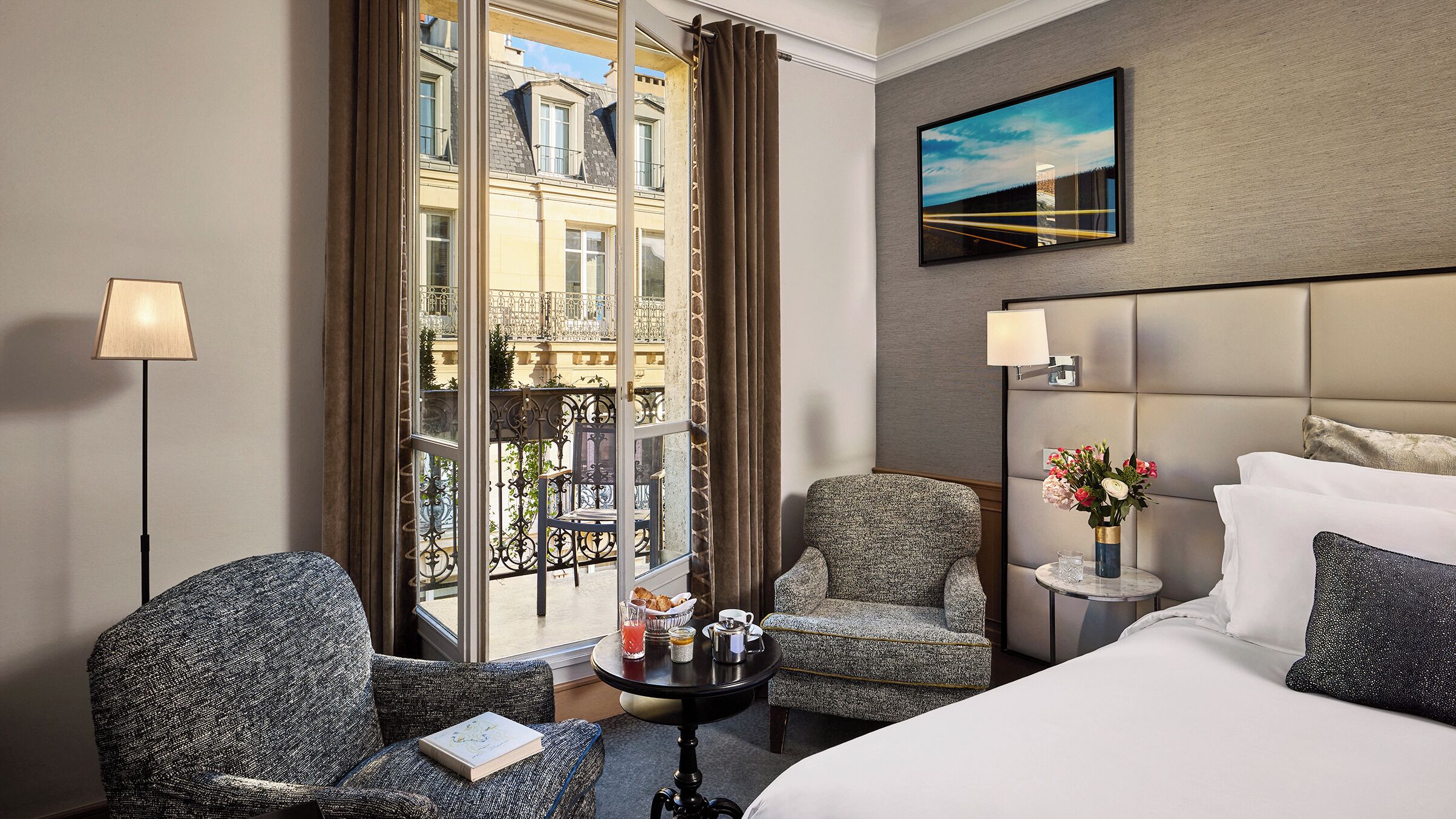 Una habitación de hotel con una cama, dos sillones, una mesita con artículos de desayuno, una mesita de noche con lámpara y vistas a un balcón con un edificio.