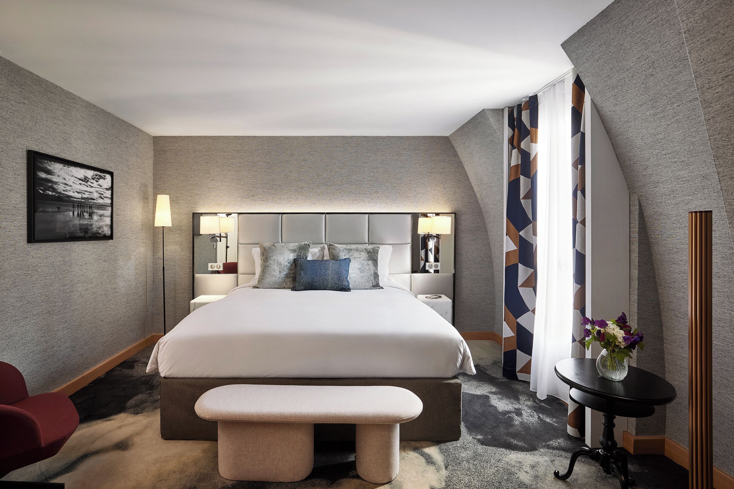 A modern hotel room with a large bed, bedside tables, a bench, and a small table with a vase of flowers.