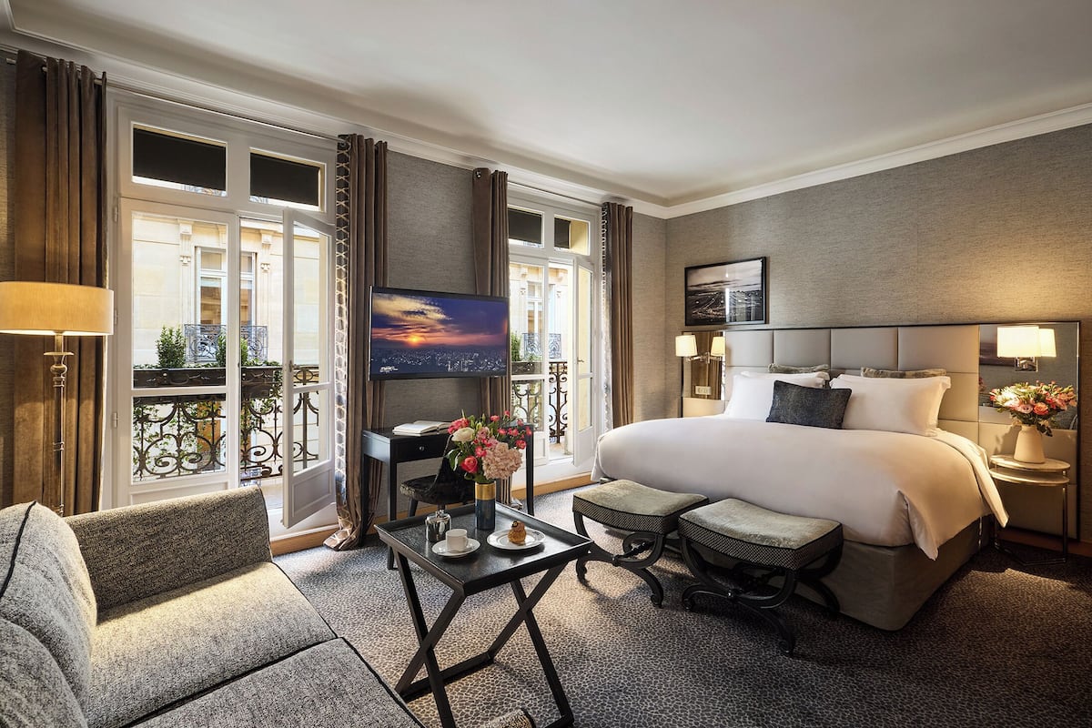 Ett hotellrum med en stor säng, en TV, ett skrivbord med en stol och en balkong med gardiner.