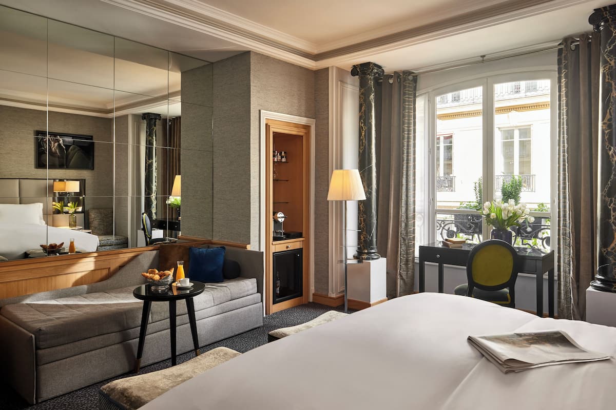 Ett hotellrum med en stor säng, en soffa, ett litet bord med en lampa, ett skrivbord och en stol. Det finns ett fönster med gardiner och utsikt över byggnaderna utanför.