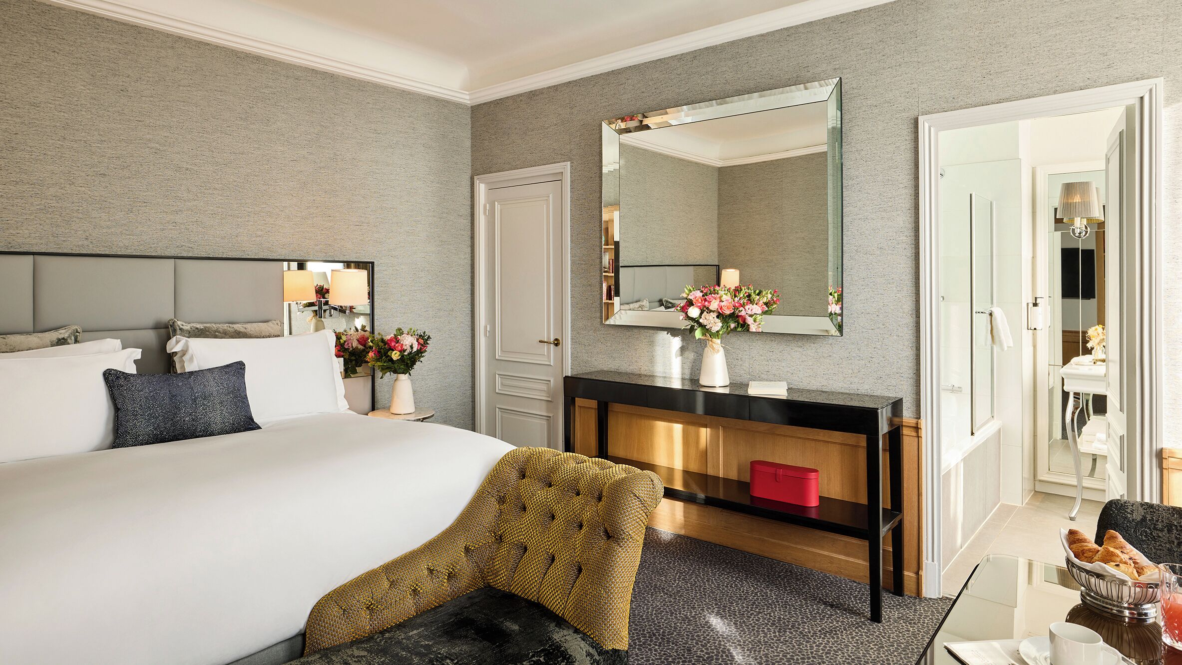 Ein Hotelzimmer mit einem großen Bett, einem Nachttisch mit Blumen in einer Vase, einem Spiegel, einem Holzkonsolentisch und einem roten Koffer.