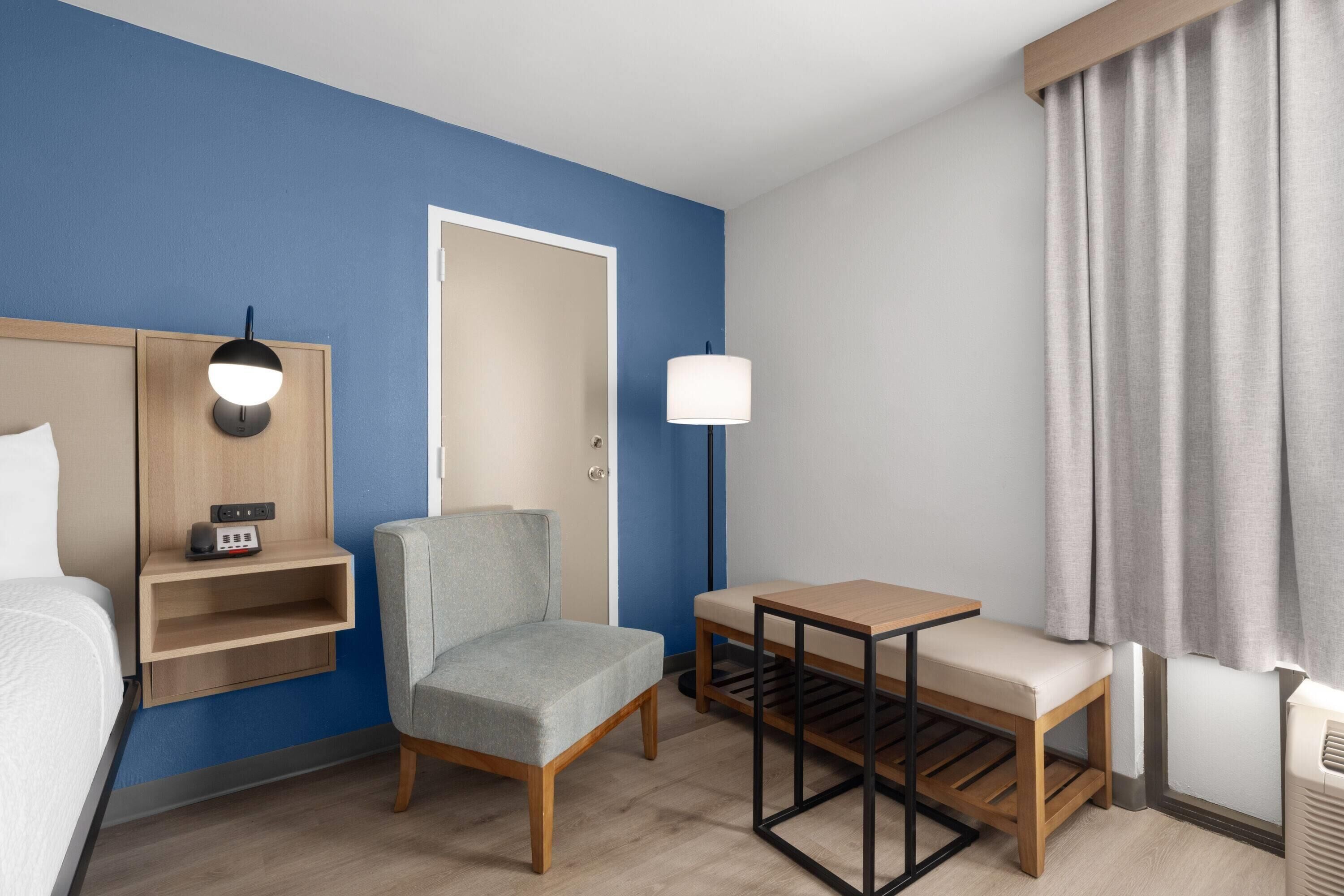 Una camera d'albergo con un letto, una sedia, un tavolino, una lampada e una finestra con tende.