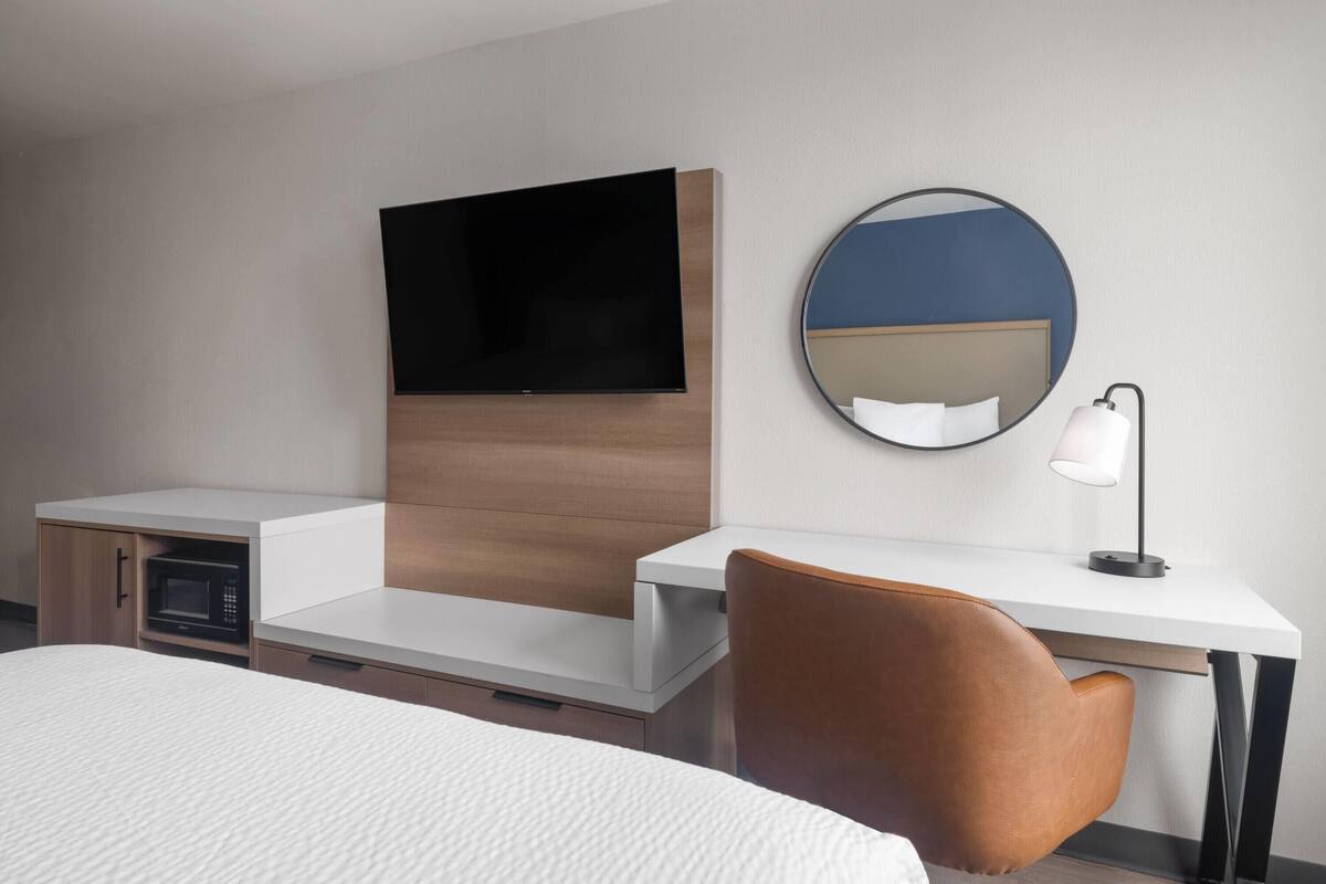 A hotel room with a bed, a desk, a chair, a television, a microwave, a mirror, and a lamp.