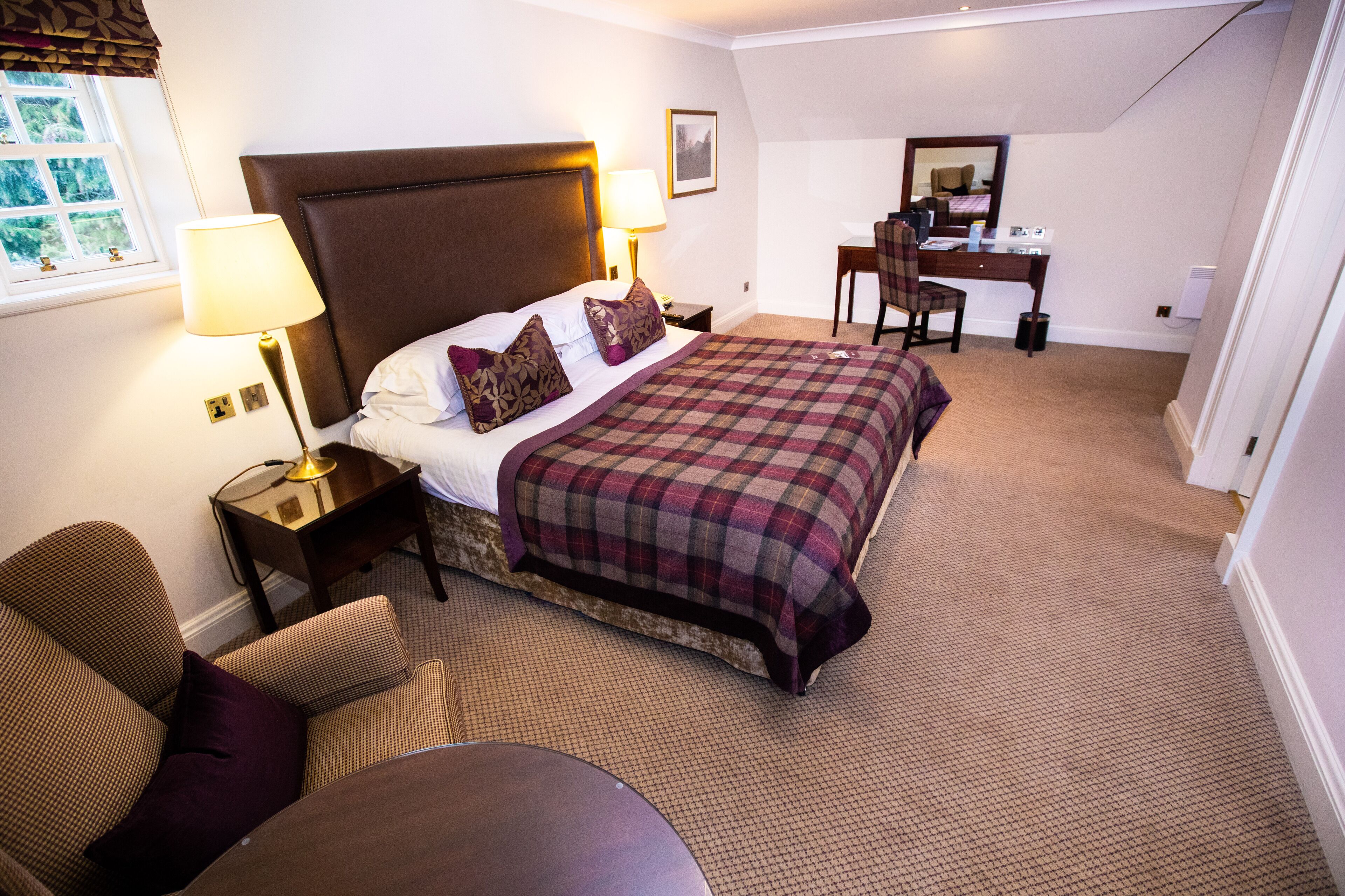 A hotel room with a large bed, two bedside lamps, a desk with a chair, and a window with curtains.