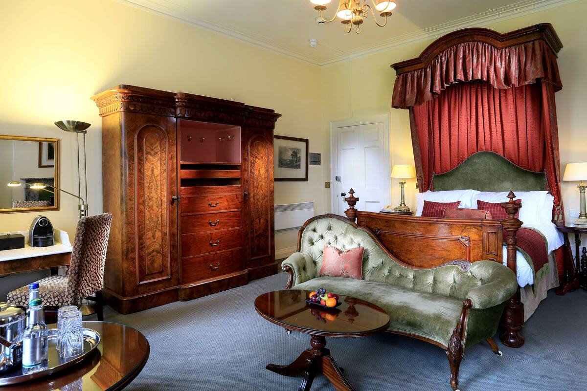 A traditional bedroom with a four-poster bed, a green armchair, a wooden wardrobe, and a small round table with a tray.