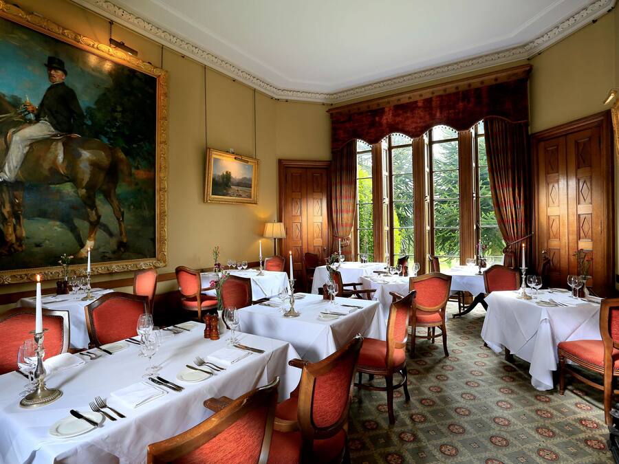 A formal dining room with tables set for service, a large painting on the wall, and a view of greenery through the windows.