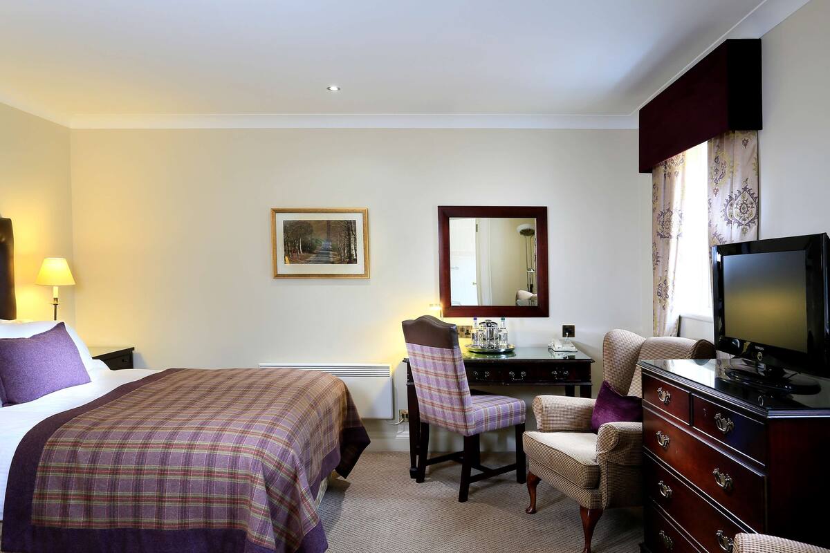 A hotel room with a bed, a desk with a chair, a dresser, a television, and a framed picture on the wall.