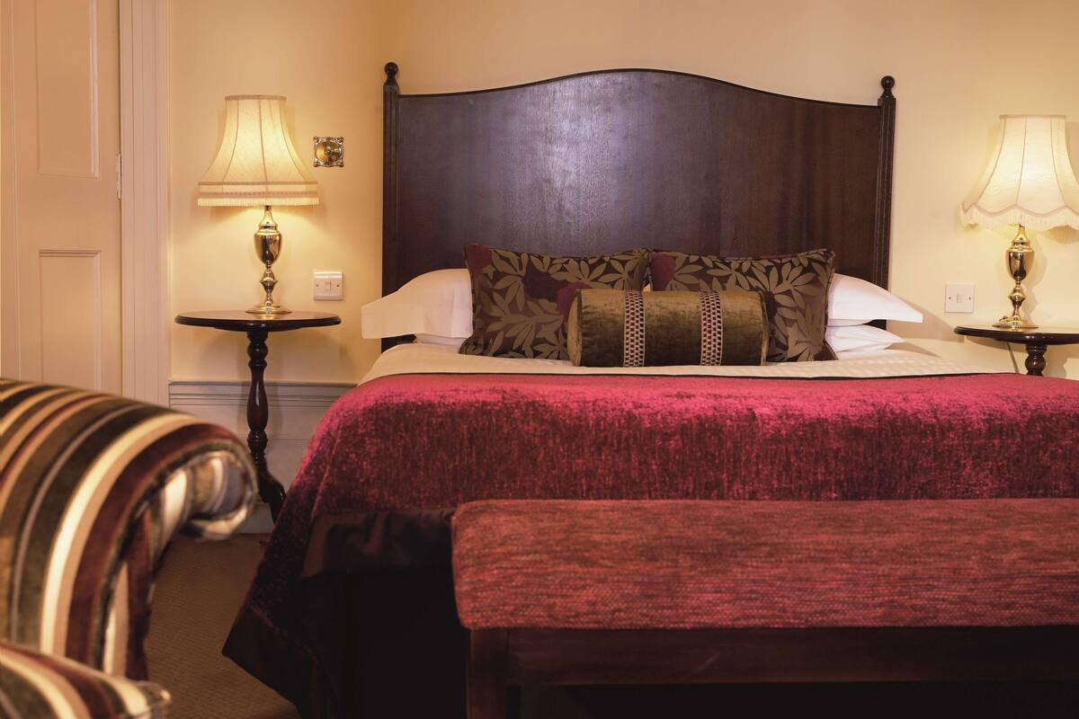 A hotel room with a large bed, two bedside tables with lamps, a striped armchair, and a bench.