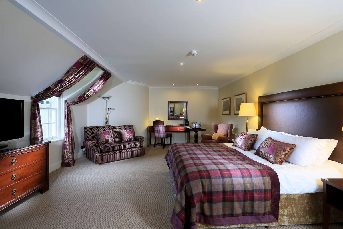 A hotel room with a large bed, a desk, two armchairs, a dresser, and a television.