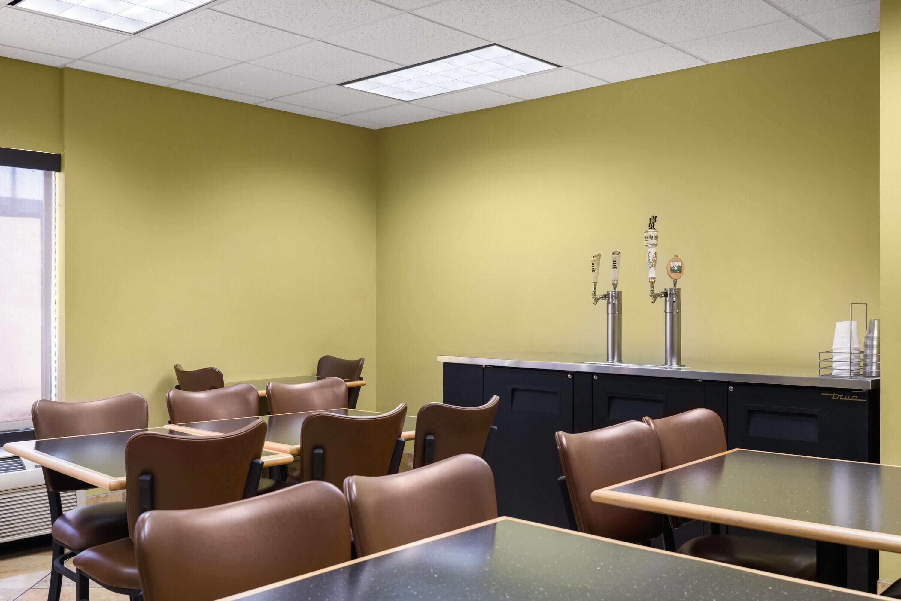 Un salón de conferencias con mesas y sillas, un mueble negro con un lavamanos y una ventana a la izquierda.