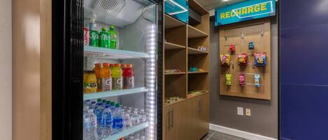 Quarto de hotel com geladeira repleta de bebidas, minibar com lanches e estação de carregamento na parede.