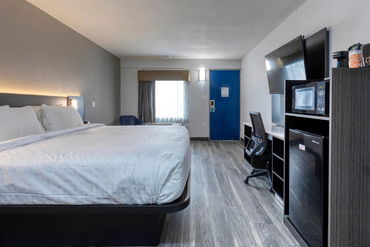 A hotel room with a bed, desk, chair, microwave, refrigerator, and a blue door.