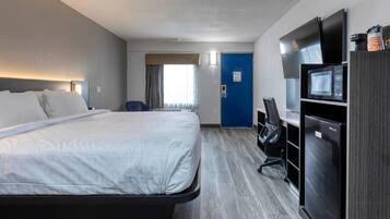 A hotel room with a bed, desk, chair, microwave, refrigerator, and a blue door.