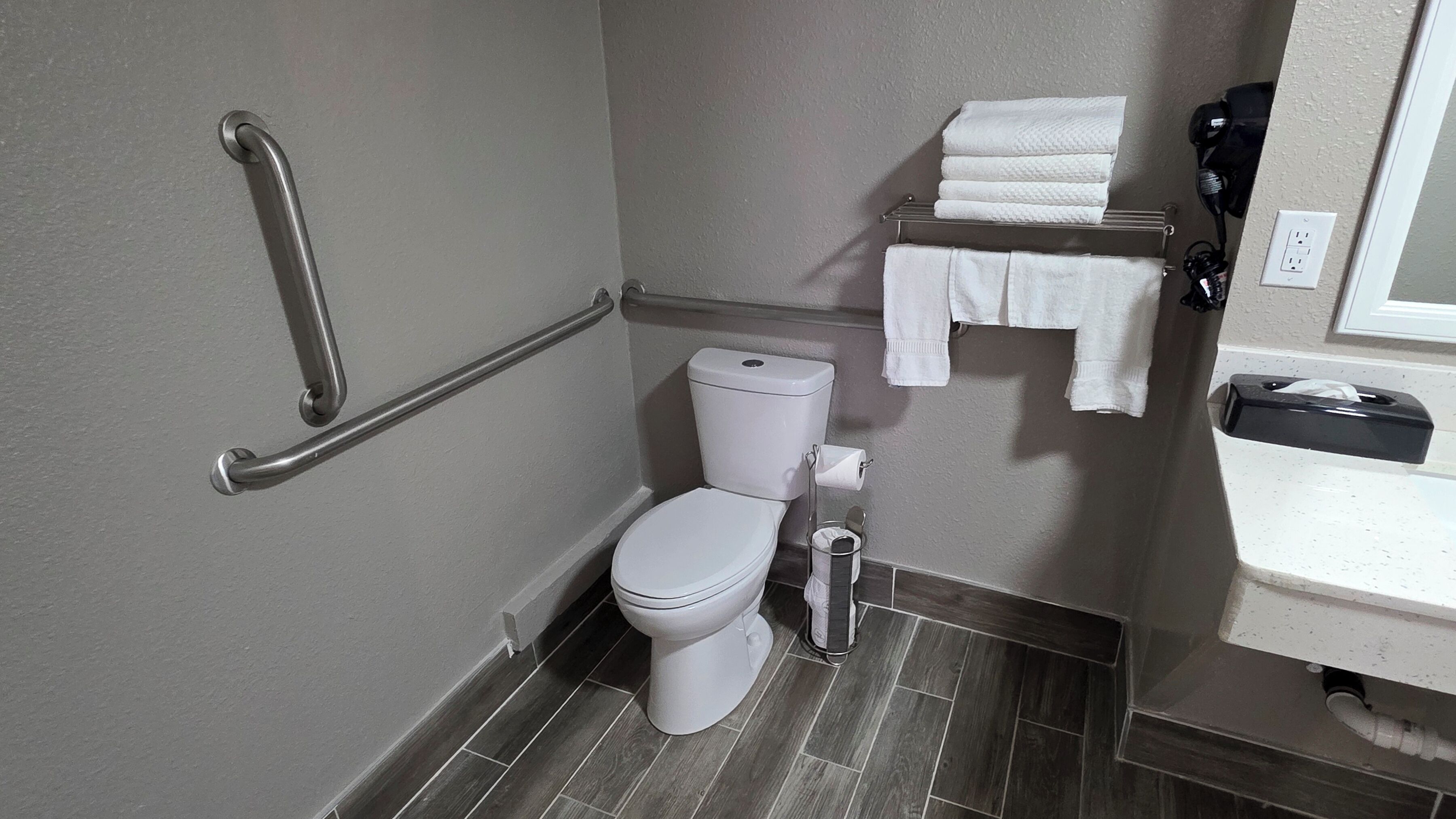 A small, modern bathroom with a white toilet, a handrail, a towel rack, and a sink with a mirror.