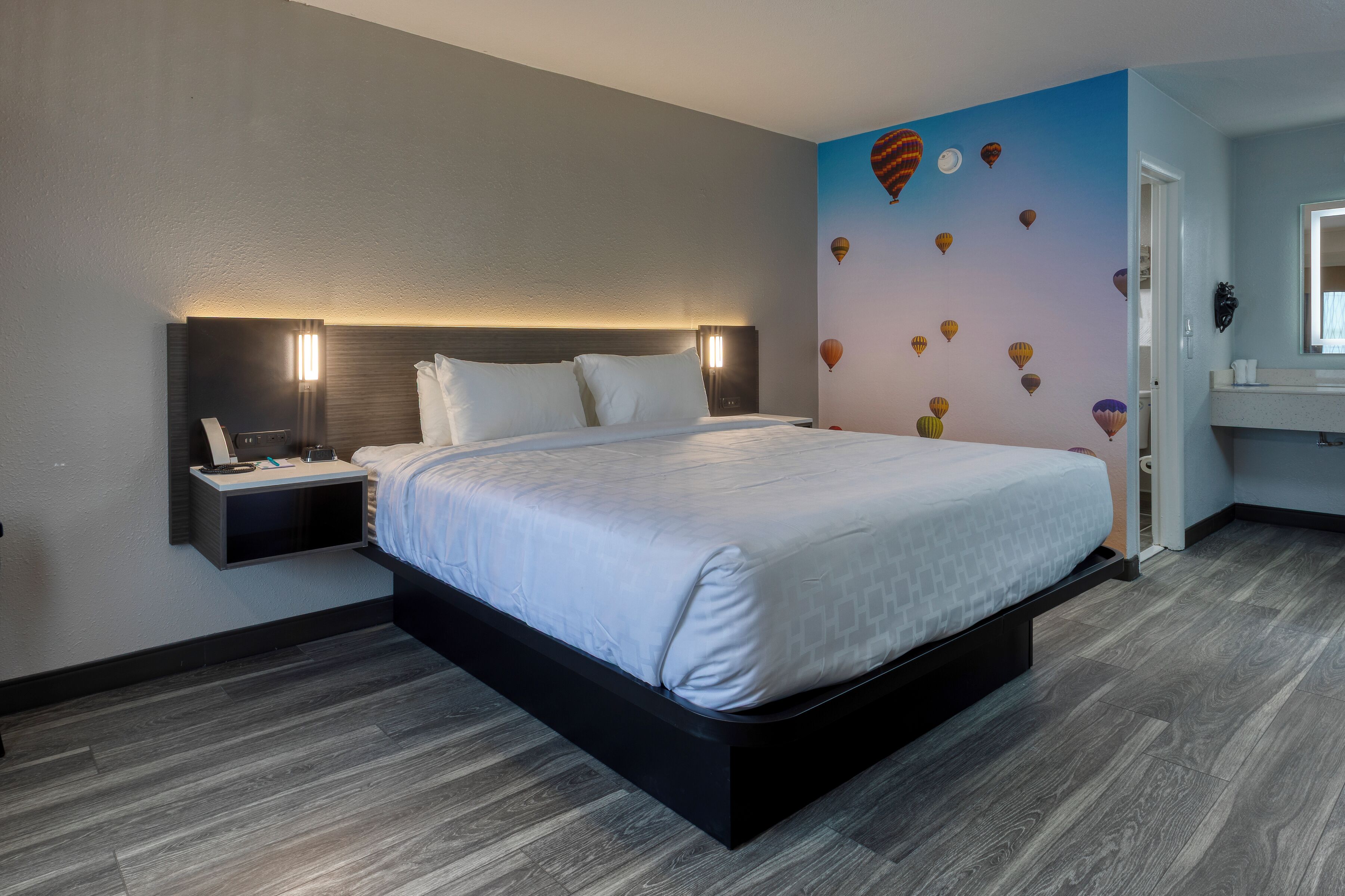 A modern hotel room with a large bed, a bedside table with a lamp, and a wall with a mural of hot air balloons.