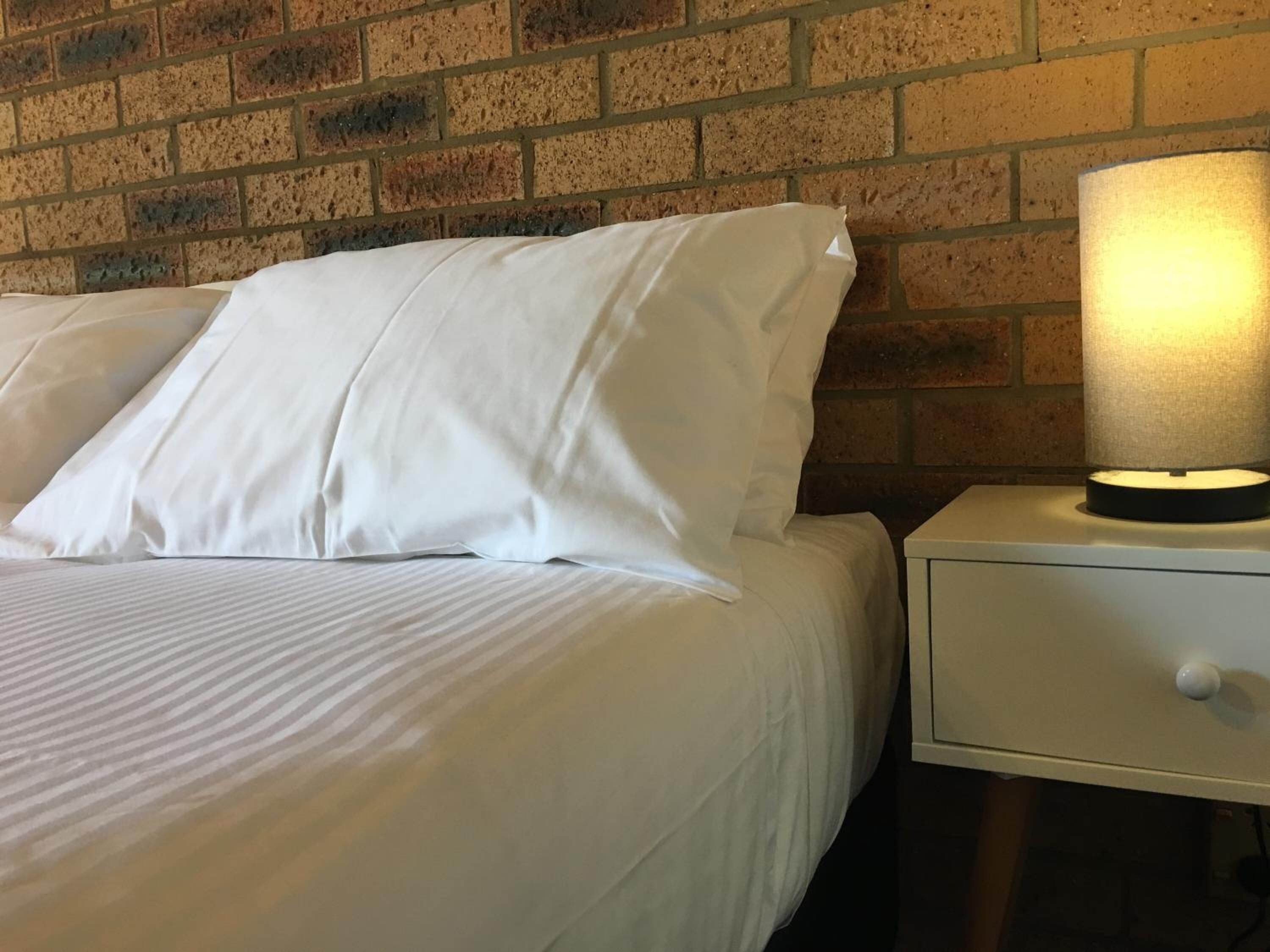 Una cama con ropa de cama y almohadas blancas, una mesita de noche con lámpara y un fondo de pared de ladrillo.
