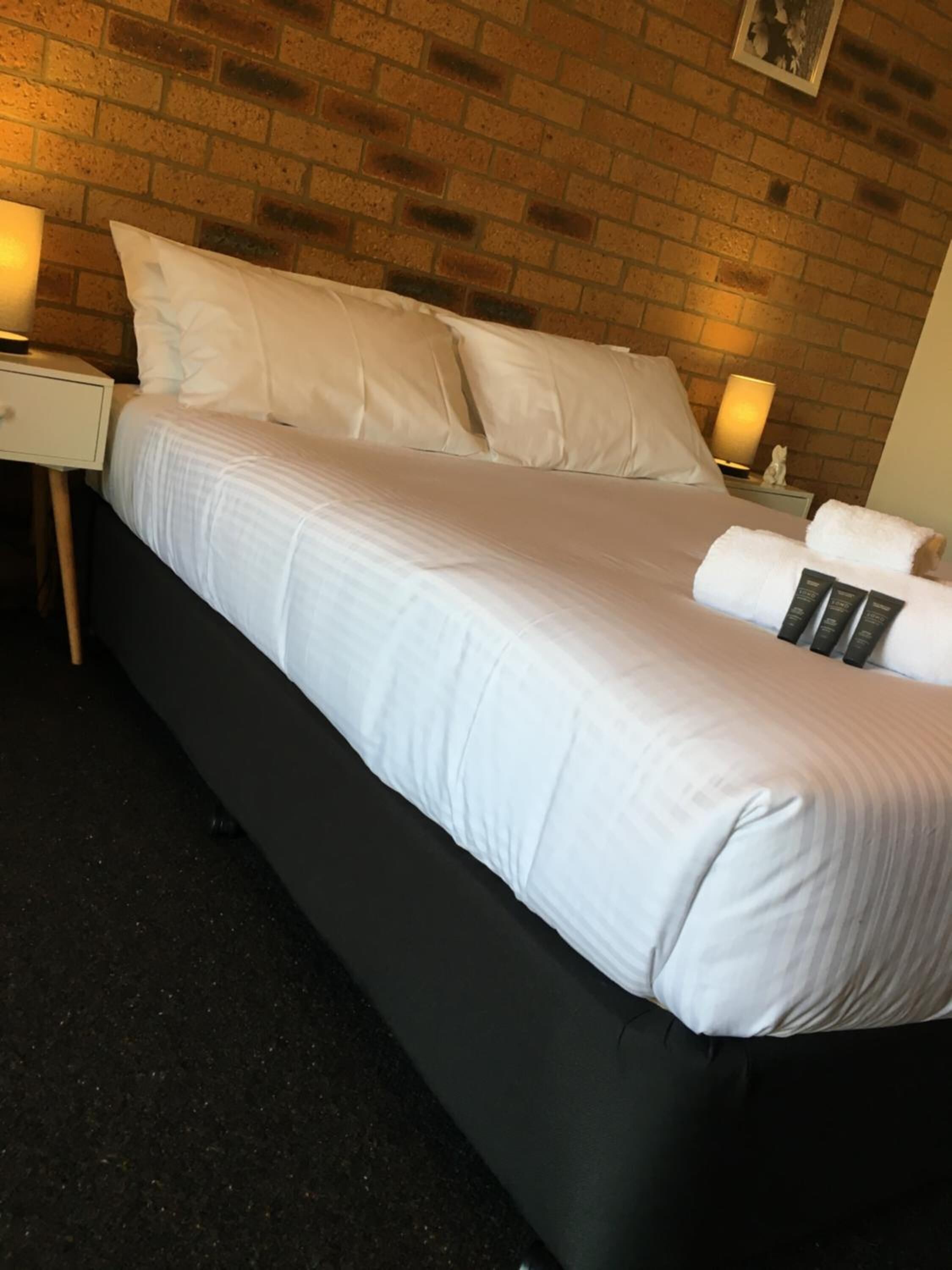 Cama bem arrumada com lençóis e almofadas brancas, cabeceira preta e parede de tijolos ao fundo.