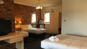 A hotel room with a brick wall, a television, two beds, a desk, and a wardrobe. - Bega Downs Motor Inn (Bega)