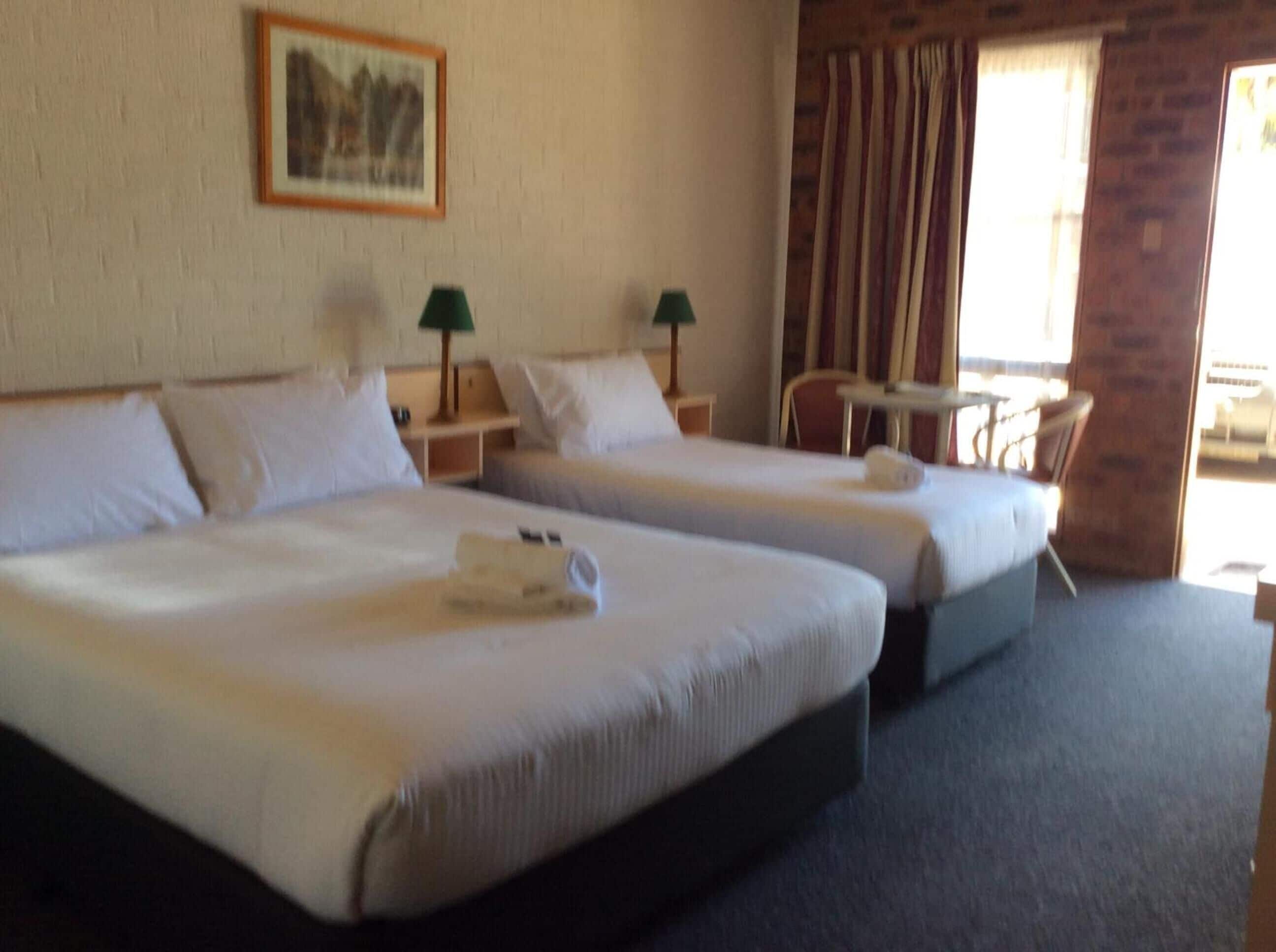 A hotel room with two beds, a small table, a chair, and a window with curtains.