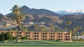 Un campo de golf con un edificio que tiene varios balcones y un paisaje montañoso de fondo.
