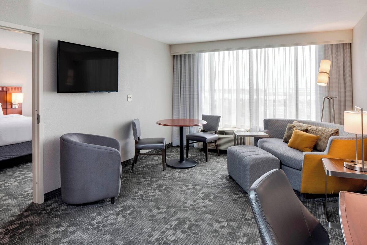 A hotel room with a flat-screen TV mounted on the wall, a gray sofa with a mustard throw pillow, a gray armchair, a small round table, and a window with sheer curtains.