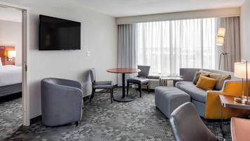 A hotel room with a flat-screen TV mounted on the wall, a gray sofa with a mustard throw pillow, a gray armchair, a small round table, and a window with sheer curtains.