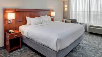 A hotel room with a large bed, two bedside lamps, a nightstand with a phone, a chair, and a large window with curtains.