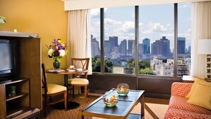A hotel room with a large window, a television, a dining table with chairs, a sofa, and a vase of flowers. - Courtyard by Marriott Boston Logan Airport (Boston)