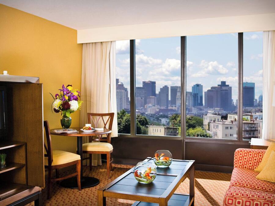 A hotel room with a large window, a television, a dining table with chairs, a sofa, and a vase of flowers.