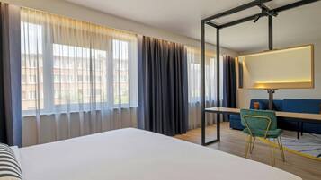 A hotel room with a large bed, a desk with a chair, a sofa, and a large window with curtains.