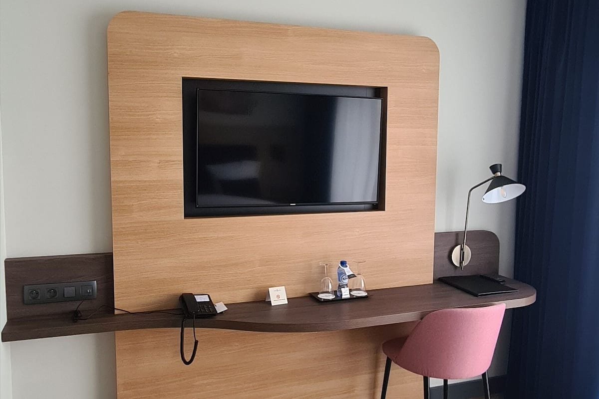 Una habitación de hotel con un escritorio de madera, un televisor de pantalla plana, una lámpara de escritorio, una silla rosa y una cama.